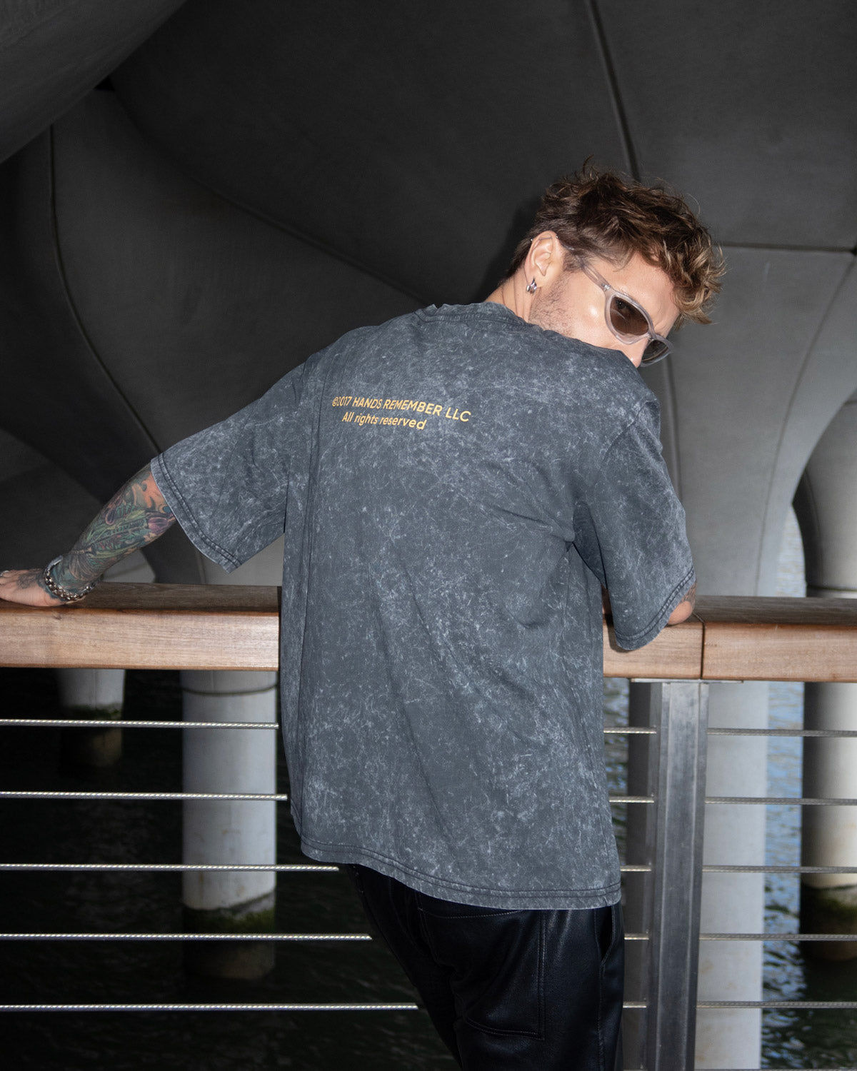 Person wearing a gray t-shirt with text on the back, standing by a railing.