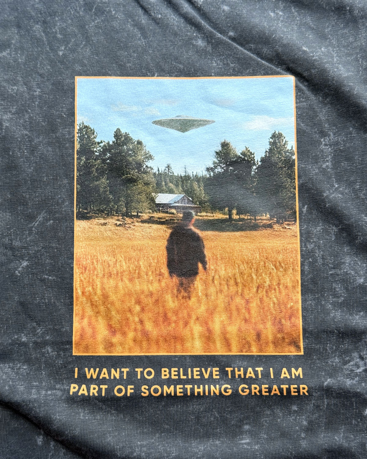 T-shirt print with a person standing in a field with a UFO and text 'I want to believe that I am part of something greater' on a textured fabric background.