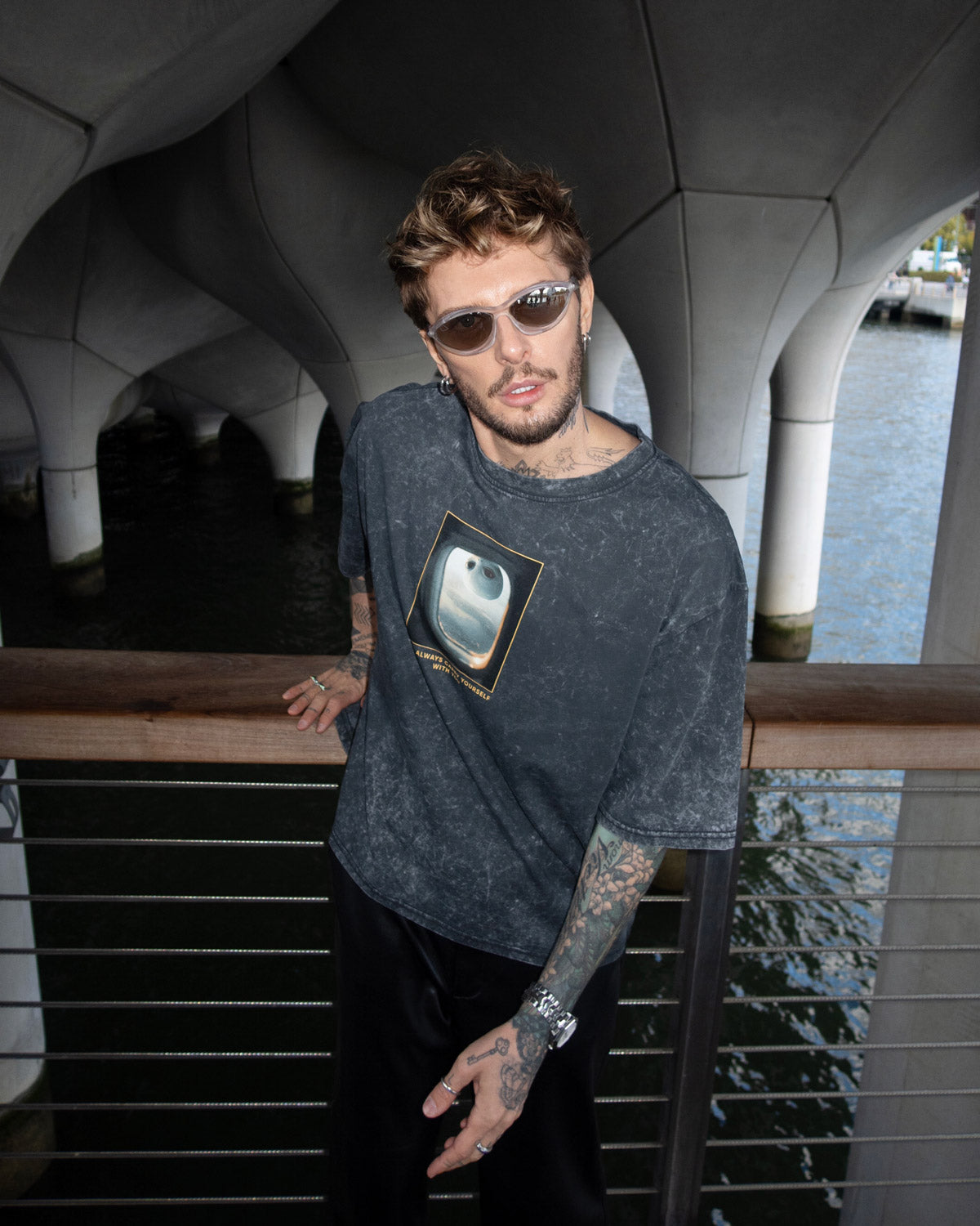 Man wearing a dark t-shirt with a graphic design, standing by a waterfront structure.