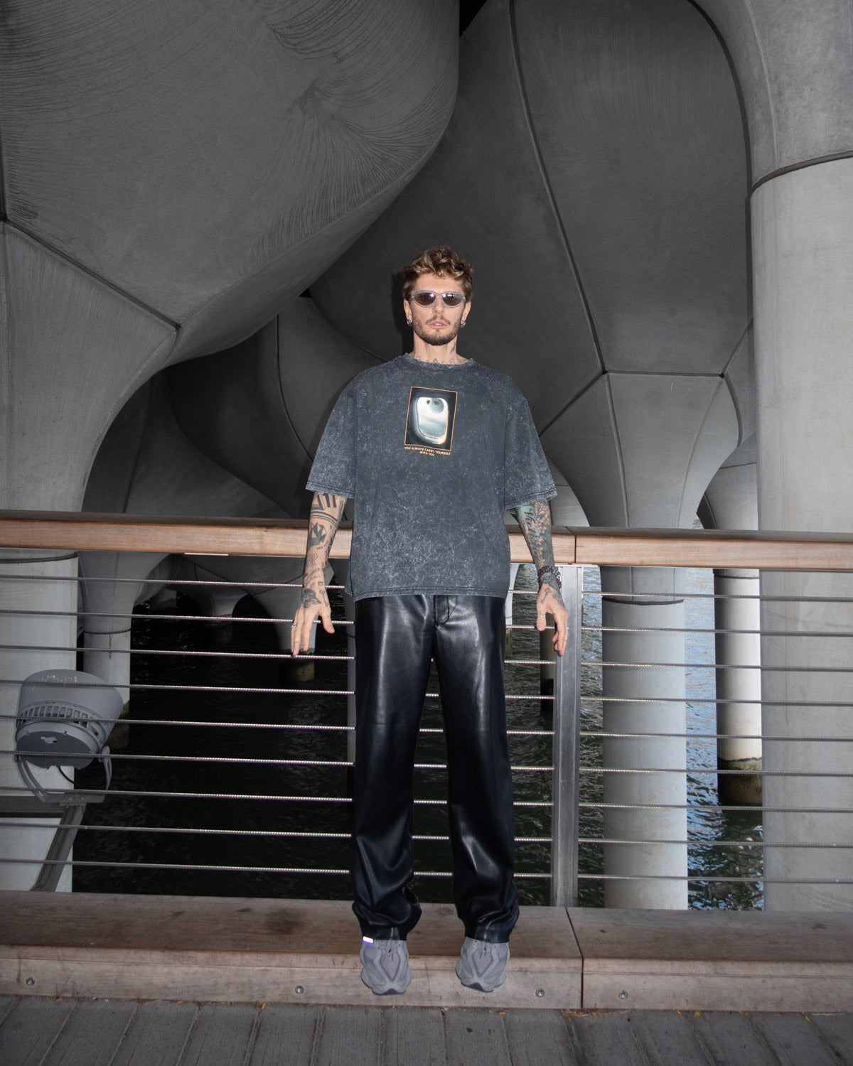 Man wearing a dark t-shirt with a graphic design, standing by a waterfront structure.