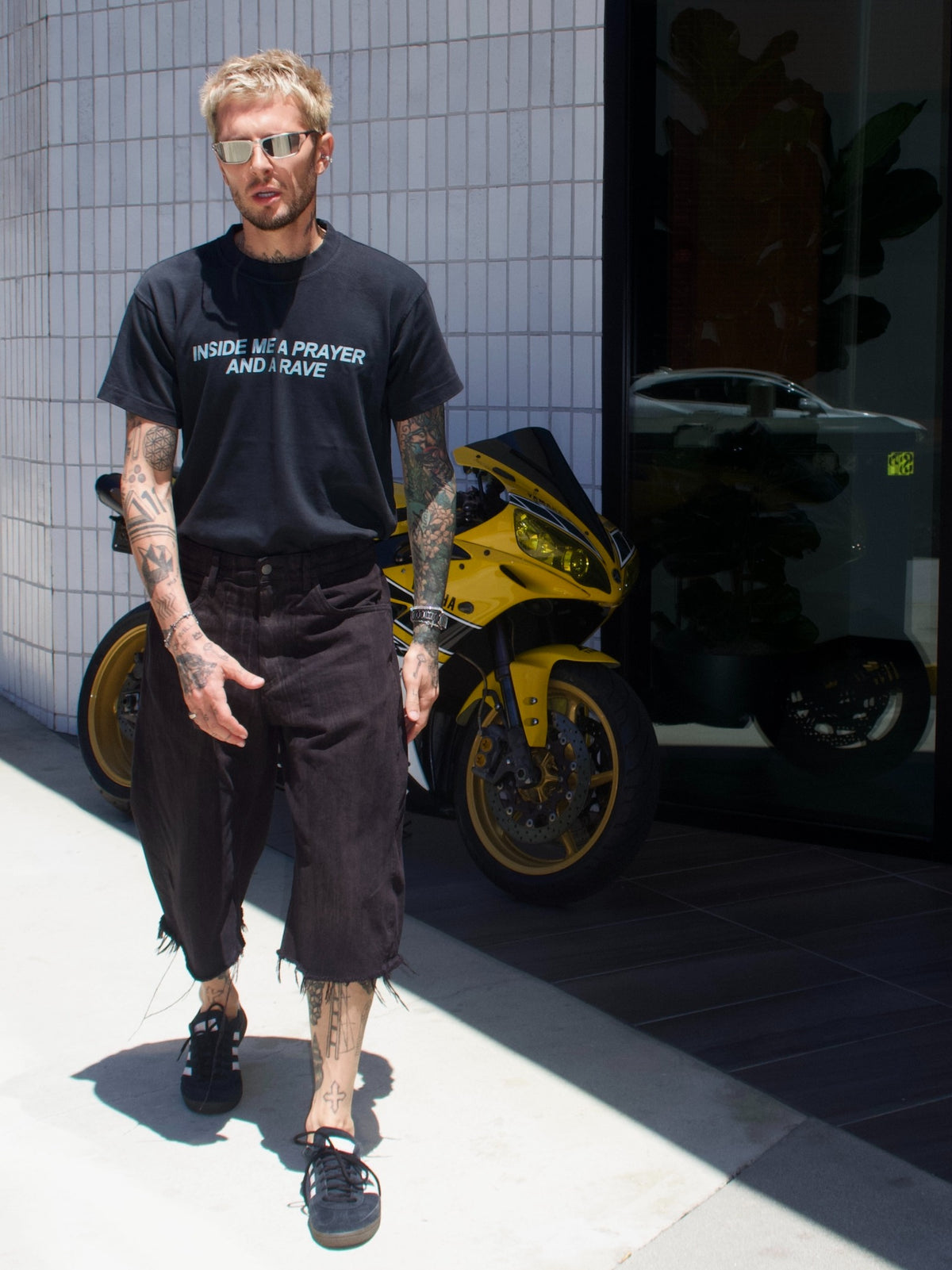 Black T-shirt with the “Inside Me a Prayer and a Rave” print, full-length front view by a yellow motorcycle