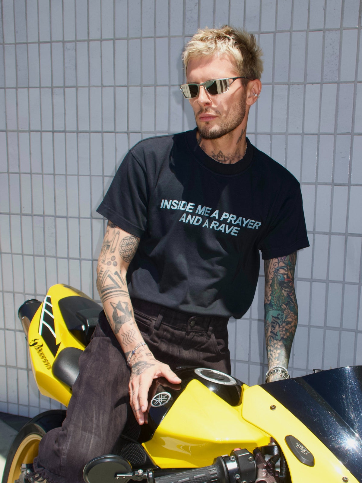Black T-shirt with the “Inside Me a Prayer and a Rave” print, seated by a yellow motorcycle