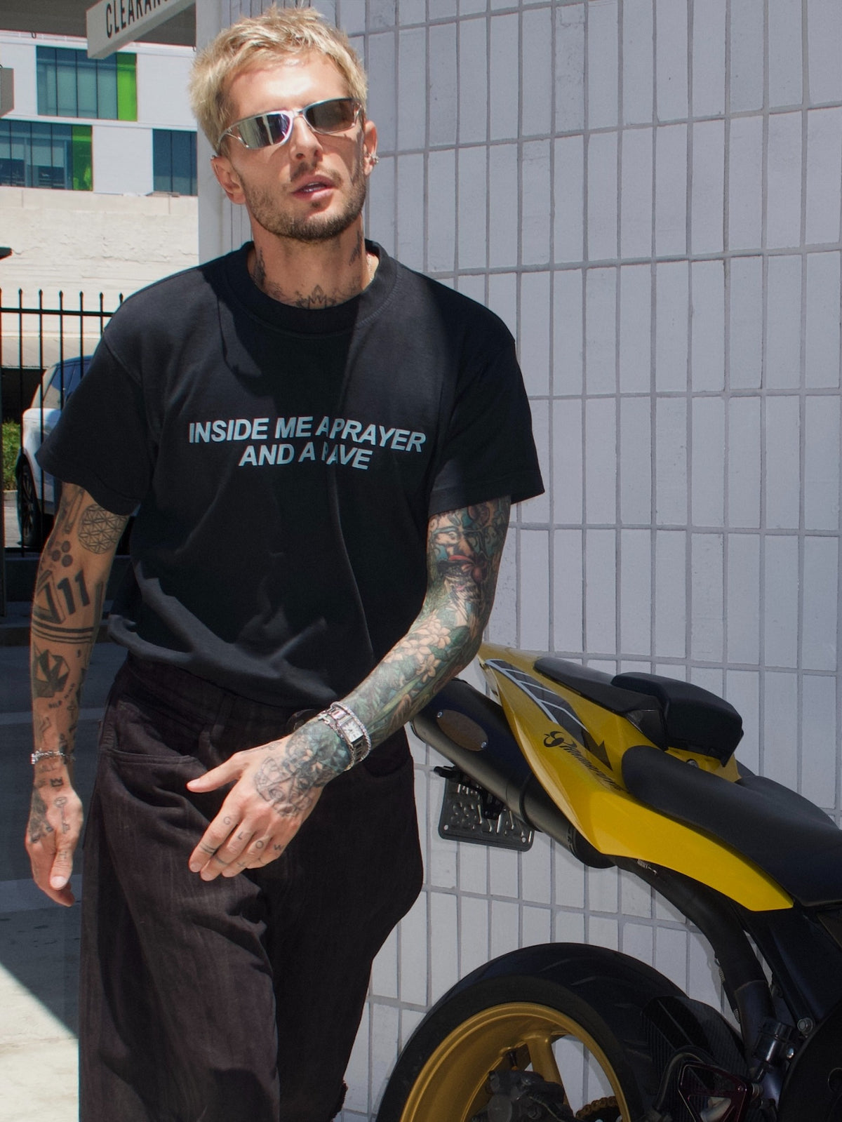 Black T-shirt with the “Inside Me a Prayer and a Rave” print, standing beside a yellow motorcycle
